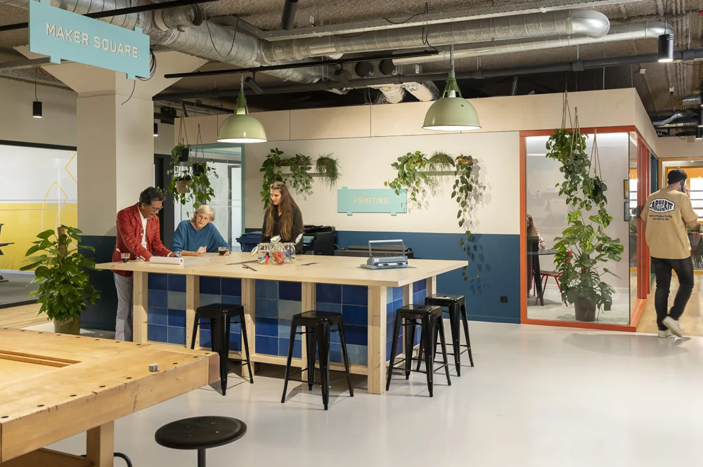 Mensen werken samen aan een lange werktafel in een creatieve ruimte genaamd Maker Square aan de Aert van Nesstraat.